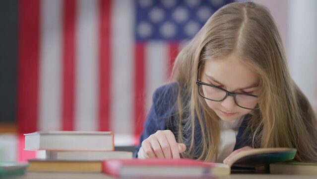 Absorbed smart teen schoolgirl reading out loud sitting on the right at desk in classroom with American flag at background. Portrait of intelligent nerd girl in eyeglasses enjoying literature