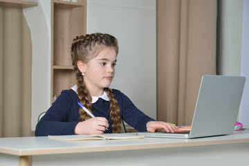 A cute school girl having online lesson using laptop and keyboard, looking at computer screen and...