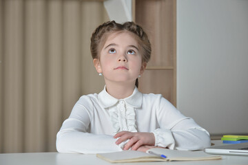 Naklejka premium A cute schoolgirl in white shirt looking up and sitting at desk. Portrait of caucasian young girl from elementary or primary school.Homeschooling, back to school and education concept.