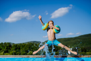 Happy kid having fun when jumping to swimming pool with inflatable armbands. Summer outdoor water activity for kids.