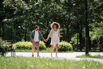 Fototapeta premium full length of happy young couple holding hands while walking together in summer park.