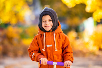 Portrait of little adorable smiling boy with scooter in beautiful autumn park with yellow, orange and green trees on sunny day