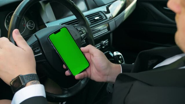 Man in business suit hands holding smartphone with vertical green screen using mobile application while driving modern car in city center.