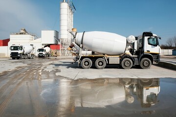 White concrete mixers standing by a modern concrete plant. Reflections of vehicles in puddle of...