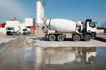 White concrete mixers standing by a modern concrete plant. Reflections of vehicles in puddle of water. © Iwona