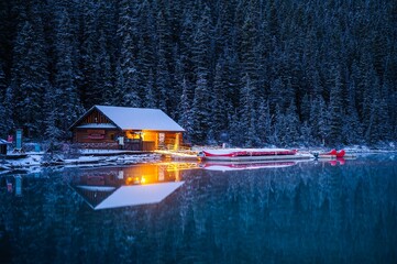Beautiful wooden cabin with lights surrounded with snow covered trees in front of a lake