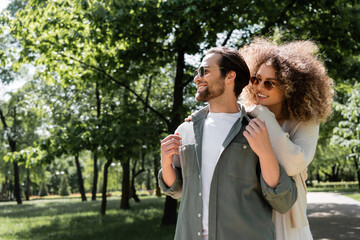 curly woman hugging happy man in stylish sunglasses in park.