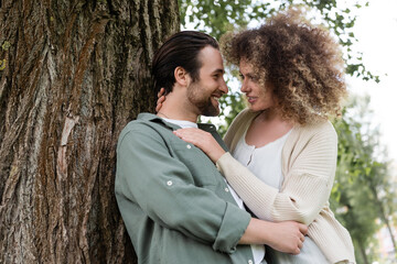 side view of young and happy couple hugging near tree trunk.