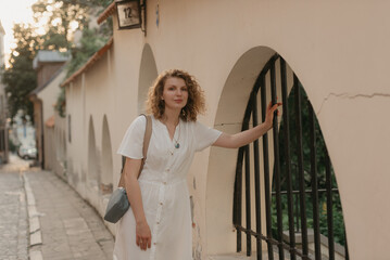 A woman with curly hair is holding on to a bar of a metal lattice in an old European town. A lady in a white dress in the evening.