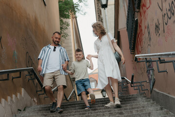 A full-length photo of a family which is going down the stairs in an old European town. A happy father, mother, and son are holding hands and having fun in the evening.