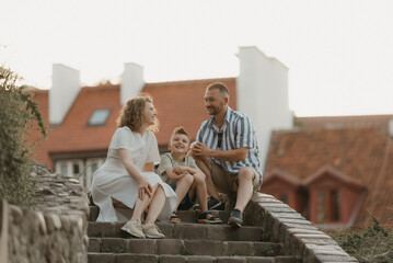 A smiling family is joking on the stairs between roofs in an old European town. A happy father, mother, and son are having fun in the evening.