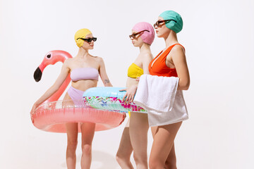 Three young women in swimming suit, cap and sunglasses posing with swimming circle isolated over grey studio background