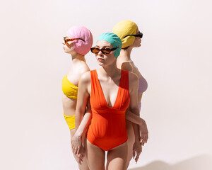 Three beautiful women in swimming suits and sunglasses standing together, posing isolated over grey studio background