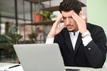 Fatigued hispanic businessman using laptop for working, the guy tired and burnout, holding head and feels headache, migraine sitting at the desk in modern office. Overwork and burnout concept