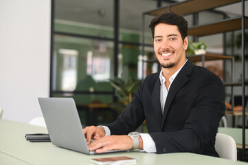 Confident latin businessman using laptop in the office. Young male office employee in formal wear typing and chatting online, develops software indoors, looks at camera with smile