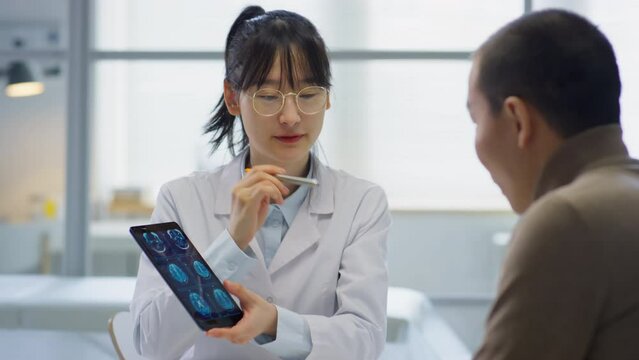 Asian female doctor showing CT scan image on digital tablet to male patient while giving medical consultation in clinic