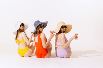 Portrait of young women sitting in swimming suit sitting, drinking cocktails isolated over grey background. Pool party
