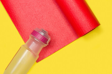 Colchoneta para yoga gimnasia roja junto a una botella de plástico sobre un fondo blanco amaillo liso y aislado. Vista superior y de cerca. Copy space. Concepto: Deportes - obrazy, fototapety, plakaty