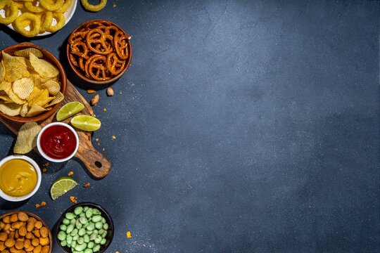 Various unhealthy salty bar snacks set. Dark grey table background with traditional party snacks - chips, onion rings, salted nuts, crisps, pretzels and sauces top view copy space