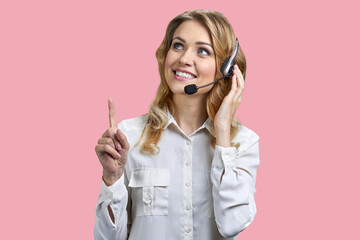 Female phone operator in headset pointing with finger upwards on pink background. Call center...