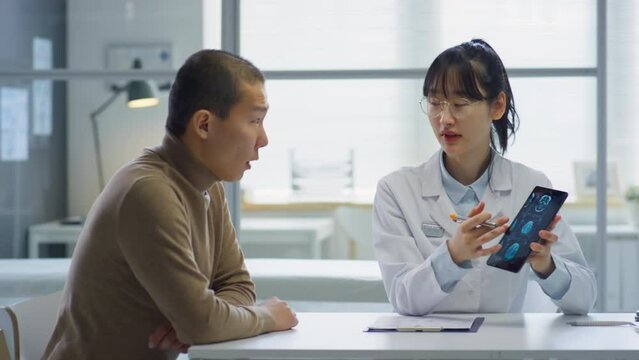 Asian female doctor explaining x-ray image on digital tablet to male patient while telling him diagnosis on medical consultation in clinic
