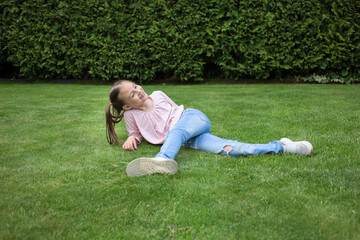 Fototapeta na wymiar Happy pretty girl 6-7 years old in jeans lies on the lawn in the city park. green nature background. Childhood outdoors, summer, spring pastime in nature