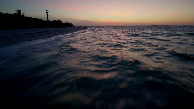 Low fast flight over the water by the shore in early dawn rising up to approach the Sanibel Island lighthouse with sinrise in the background
