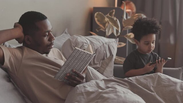 Slowmo of Black man lying in bed and reading book while his toddle son playing games on smartphone sitting nearby, spending evening leisure time together