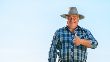 Delighted mature male worker looking at camera with thumb up gesture while standing in agricultural plantation on summer day in rural area. Smiling farmer showing thumb up gesture in field copy space © Alexandr