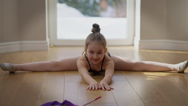 Front view portrait of confident motivated teenage gymnast looking at camera bending forward on twine smiling. Happy inspired Caucasian teenage girl posing working out stretching muscles indoors