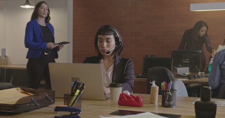 Office place with young people working at corporate job. Female staff wearing headset behind laptop...
