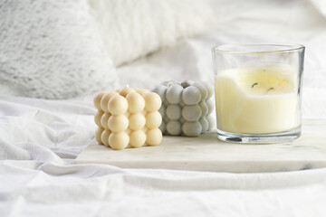 Three candles - bubble candles and white candle in glass jar with silver lid on marble tablet on white bed sheets with grey fluffy pillows
