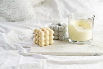 Three candles - bubble candles and white candle in glass jar with silver lid on marble tablet on white bed sheets with grey fluffy pillows