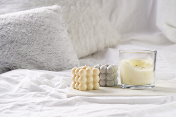Fototapeta na wymiar Three candles - bubble candles and white candle in glass jar with silver lid on marble tablet on white bed sheets with grey fluffy pillows