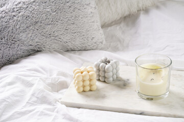 Three candles - bubble candles and white candle in glass jar with silver lid on marble tablet on white bed sheets with grey fluffy pillows