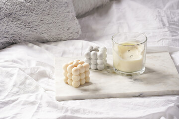 Three candles - bubble candles and white candle in glass jar with silver lid on marble tablet on white bed sheets with grey fluffy pillows