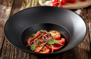 Traditional uzbek salad with tomatoes and sweet onion on black bowl. Tomatoes and onion salad on wooden background in rustic style. Appetizer from tomato and coriander.