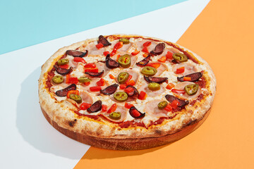Italian pizza with sausage, jalapeno, ham and paprika on coloured background. Spicy pizza with jalapeno in minimal style on blue and orange color. American pizza delivery concept with color backdrop.