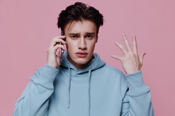 a handsome, thoughtful, puzzled man stands on a pink background in a light blue hoodie and holding a smartphone in his hands talks on it, actively gesticulating with his hand. 