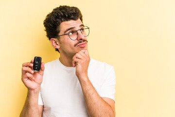 Young caucasian man holding car keys isolated on yellow background looking sideways with doubtful...