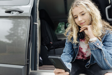 Fototapeta na wymiar Young teenage girl sitting in camping van and looking at her cell phone