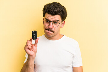 Young caucasian man holding car keys isolated on yellow background confused, feels doubtful and unsure. - obrazy, fototapety, plakaty