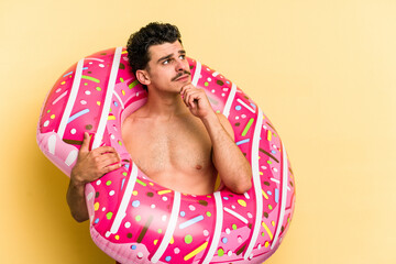 Young caucasian man holding an inflatable donut isolated on yellow background looking sideways with...