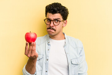 Young caucasian man holding an apple isolated on yellow background confused, feels doubtful and...
