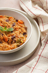 Grain dish - Millet with tomatoes, beans, carrots and spices, mint in a white bowl on a concrete round tray with checkered linen kitchen towel on purple background