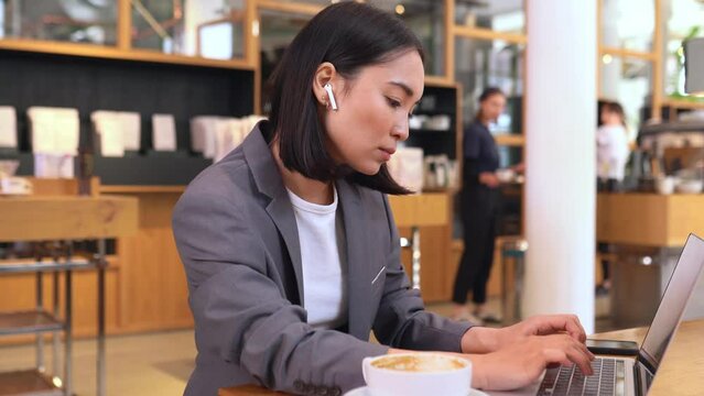 Busy smart young Asian professional business woman using laptop computer drinking coffee during lunch in cafe. Korean lady entrepreneur or manager remote distance working online watching webinars.