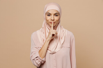 Young arabian asian muslim woman she wear hijab pink clothes say be quiet, finger on lips hush gesture isolated on plain pastel light beige background People uae middle eastern islam religious concept