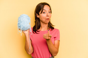 Young hispanic woman holding a ball of wool isolated on yellow background pointing with finger at you as if inviting come closer.