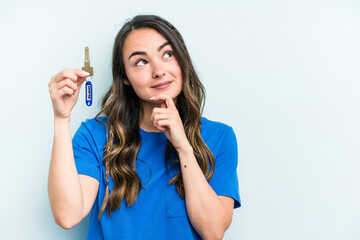 Young caucasian woman holding home keys isolated on blue background looking sideways with doubtful...