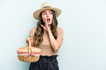 Young caucasian woman holding a picnic basket isolated on blue background is saying a secret hot...